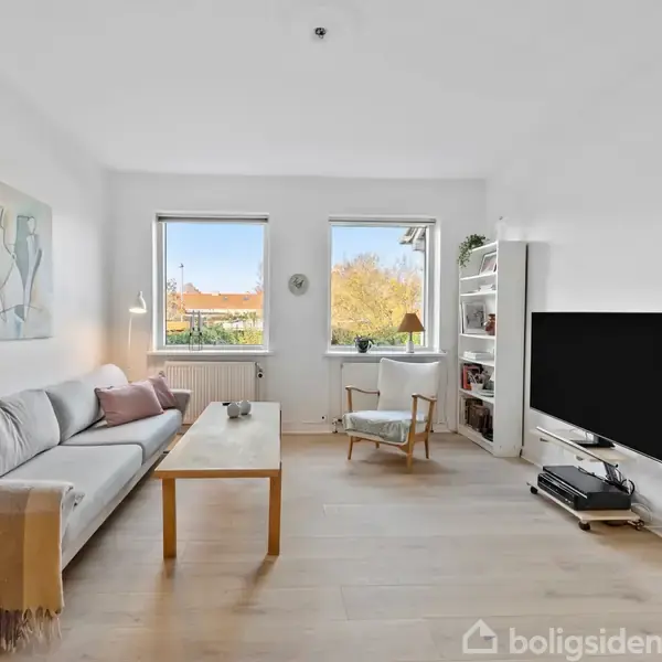 En stue med en sofa til venstre, et bord i midten, en stol ved vinduet og en fladskærms-tv til højre, omgivet af hvide vægge og træmøbler.