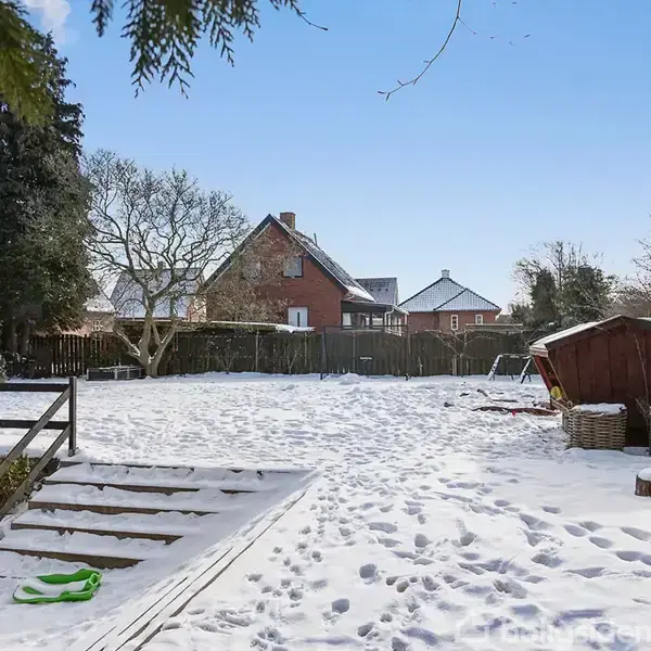 Sneklædt have med træstubbe i nederste højre hjørne; baggrunden viser et rødt murstenshus og andre bygninger omkranset af træer. Trætrapper fører op til det sneede område.
