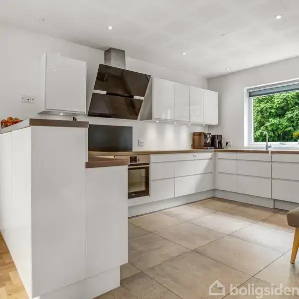 Et moderne køkken med hvide skabe og integreret ovn. Rummet er lyst med et stort vindue med udsigt til grønne træer. En grå bænk står ved siden af et spisebord. En vægur hænger på væggen.