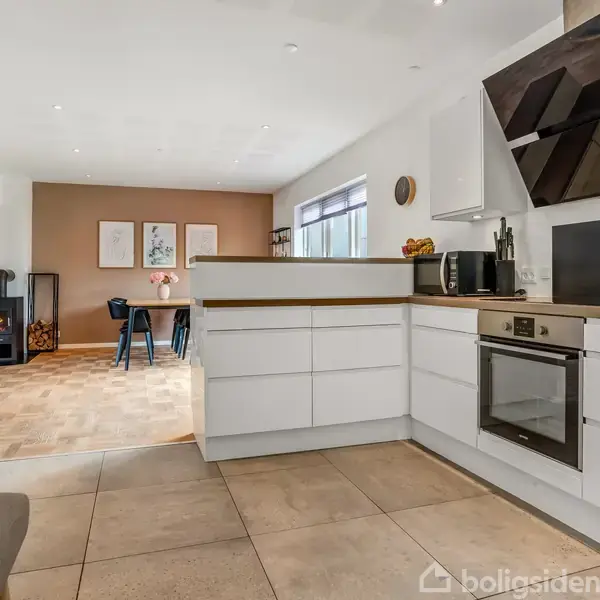 Moderne køkken med hvide skabe og indbygget ovn, der åbner ud mod en stue. Stuen har spisebord og en brændeovn i hjørnet, med lyst trægulv og kunst på væggen.