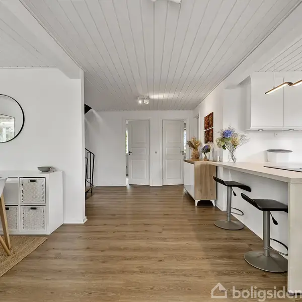 Spisebord med fire stole står til venstre. Køkkenø med to barstole til højre. Lys, moderne indretning med træpanelgulv, hvidmalede vægge og loft samt dekorative elementer som planter og spejl.