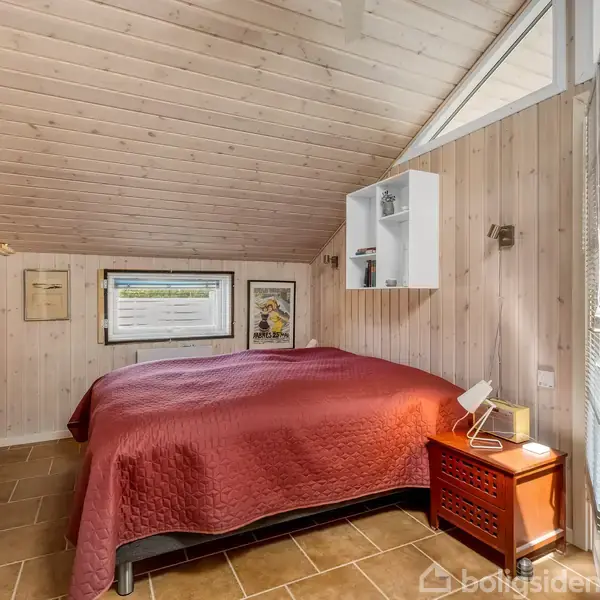 En seng med rød sengetæppe står i et lyst soveværelse med trævægge og loft. Ved siden af hænger der en fladskærms-TV på væggen, og der er en udgang til terrasse.