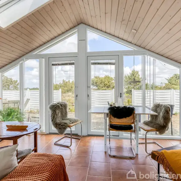 Stue med glasdøre og store vinduer slipper naturligt lys ind, med et åbent spisebord og stole dækket af lammeskind. Lyst træloft og flisegulv skaber en hyggelig atmosfære.