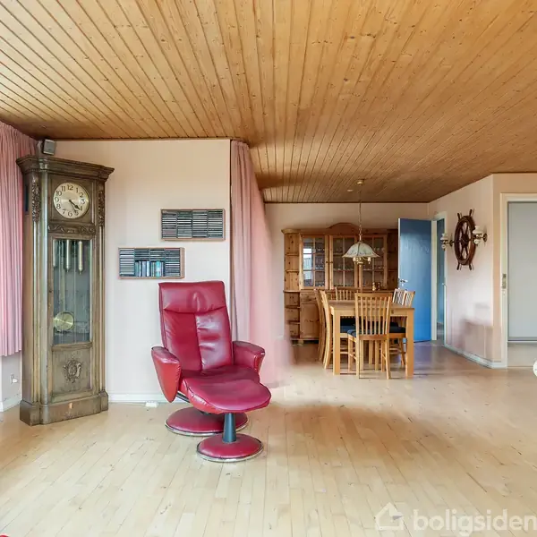 Stue med rød lænestol og gulv-til-loft ur til venstre. I baggrunden ses et spisebord med stole og en vitrineskab. Rummet har trægulv og loft, med store vinduer til venstre.