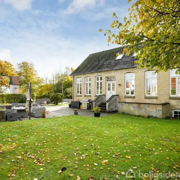 Et murstenshus med sorte tagsten står i midten, omringet af en grøn græsplæne og efterårsfarvede træer. På terrassen foran huset er der havemøbler og parasoller.