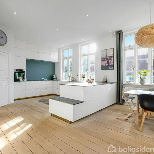 Stue-køkken med hvide skabe og køkkenø, dagslys ind gennem store vinduer, spisebord i højre hjørne med sorte og træstole, vægdekorationer og en gulvplante tilføjer farve.