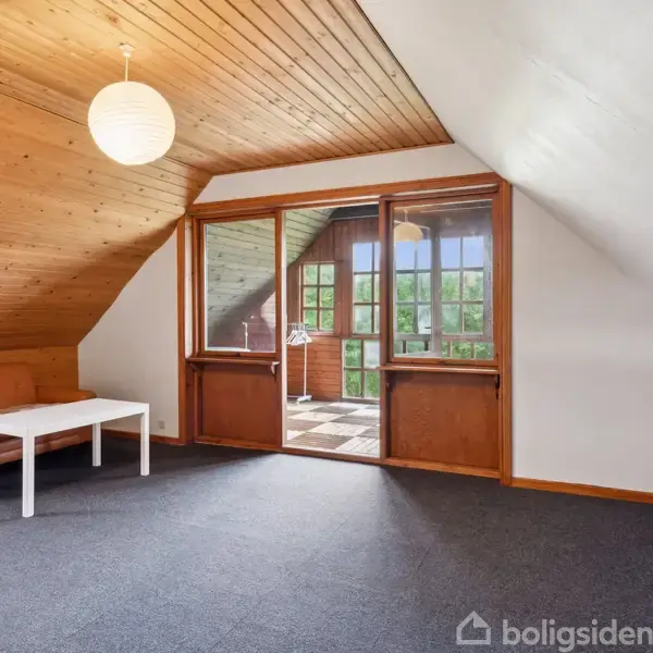 En stue med en brun lædersofa og hvidt sofabord placeret på et mørkt tæppe. Store vinduer med udsigt til en overdækket balkon. Træloft og hængende lampe.