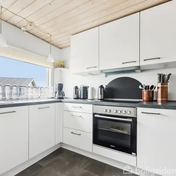Hvidt køkken med sorte bordplader, hvor skuffer og skabe er lukkede. Kaffemaskine, elkedler og knivblok står på bordet. Store vinduer giver udsigt til tag og blåt himmel.