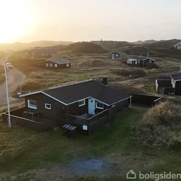 Et fritliggende sommerhus står stille i et åbent, bakket landskab ved solnedgang. Flagstang og nabohuse ses omkring, og det er omgivet af græs og stier.