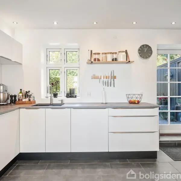 Et lyst køkken med hvide skabe og en sort bordplade. Et vindue og en dør lukker lys ind. Over bordpladen hænger køkkenredskaber, en hylde med krukker og et ur.