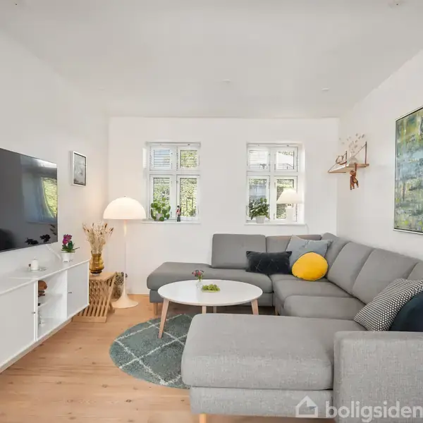 Grå hjørnesofa med farverige puder står i et lyst stueområde med trægulv. Væghængt TV og reol til venstre, dekorative planter og kunst på væggen.