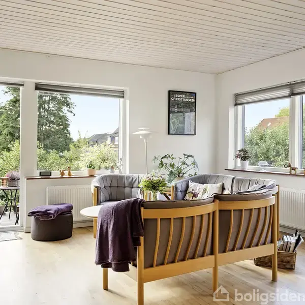 En lys stue med træmøbler og grå puder står over for store vinduer, som giver udsigt til en grøn have og terrassen. En plakat hænger på væggen, og en radiator ses under vinduet.