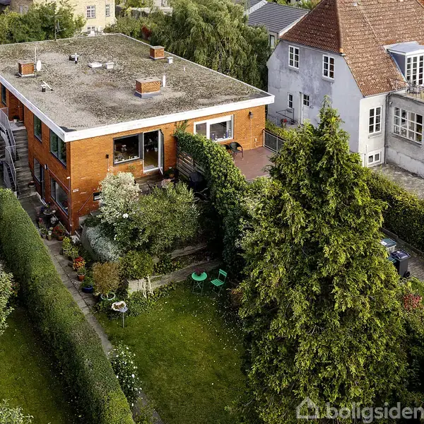 En fleretagers murstensbygning med fladt tag i en beboelsesområde. Den er omgivet af en grøn have med buske og træer samt en terrasse. Naboens hus ses til højre.