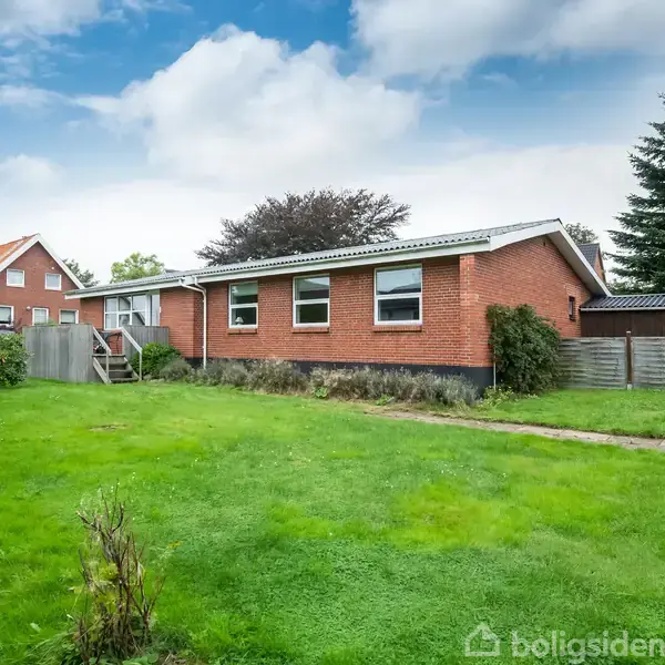 Rødt murstenshus står stille i en stor have med græsplæne, omgivet af buske og træer. En anden bolig ses i baggrunden under en nuanceret blå og skyet himmel.