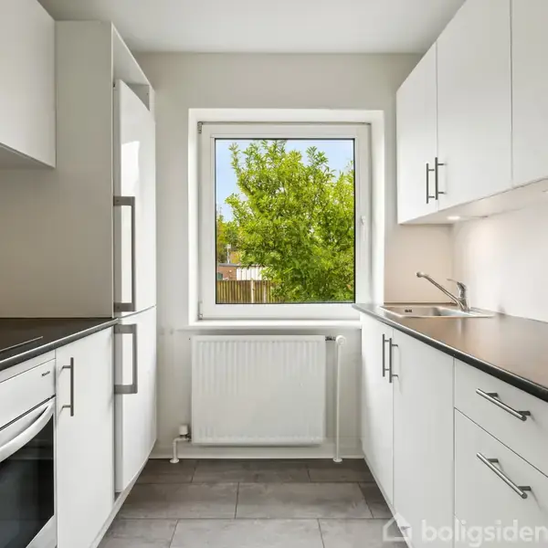 Hvidt køkken med indbyggede skabe og skuffer åbner mod et vindue, der viser et grønt, frodigt træ udenfor. Grå fliser på gulvet og et moderne komfur ses.