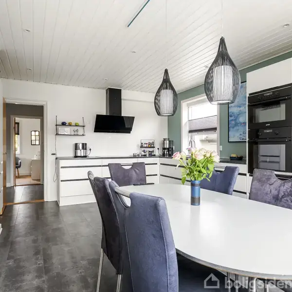 Et moderne køkken med et ovalt, hvidt spisebord omgivet af blå stole. Bordet har en vase med blomster. Sorte lamper hænger over bordet. Køkkenet har hvide skabe og en sort emhætte.