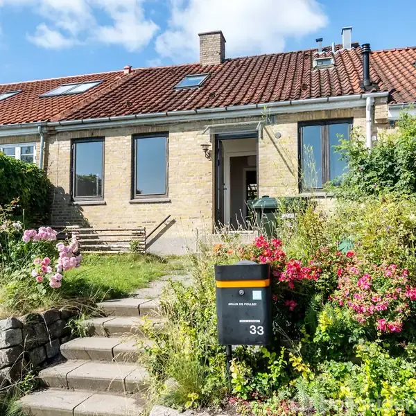En rækkehusfacade i gule mursten med to store vinduer. Foran huset er en lille have med lyserøde blomster, og en trappe fører op til hoveddøren.