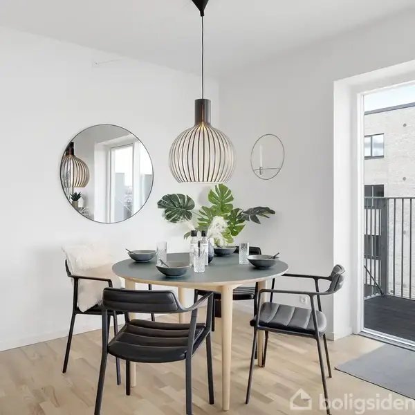 Rundt spisebord med fire sorte stole står i en lys spisestue. Bordet er dækket med tallerkener og planter. En rund vægspejl hænger over bordet ved siden af en stor glasdør til en altan.