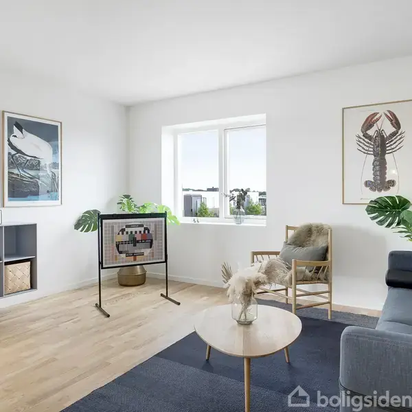 Stue med gråt hjørnesofa står på et blåt tæppe. Et rundt sofabord har en vase med tørrede blomster. Væggene har kunst, og et vindue giver naturligt lys.