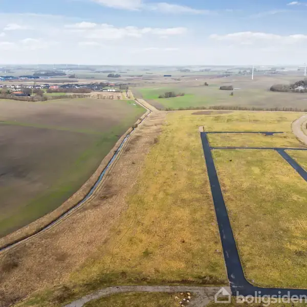 Et åbent landområde med marker dominerer udsynet. Asfalterede stier løber gennem landskabet, og vindmøller er synlige i baggrunden under en blå himmel med spredte skyer.