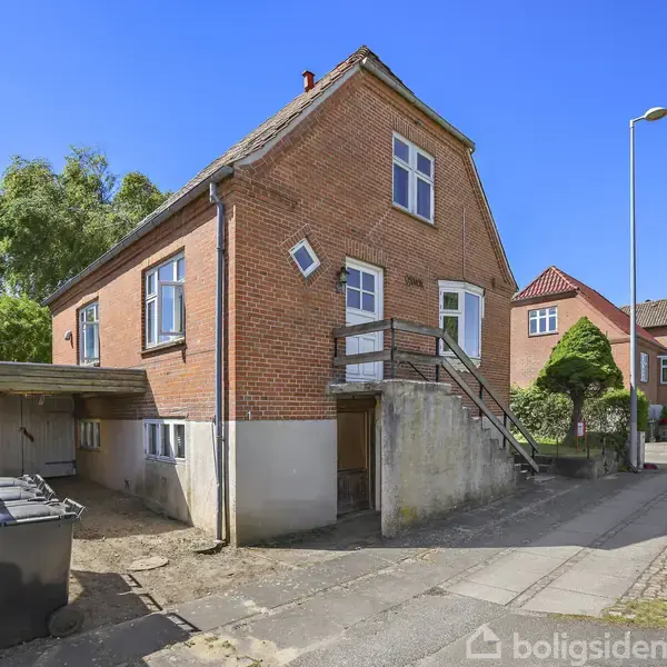 Rød murstenshus med to etager, trappe til hoveddør, beliggende på en stille gade. Omkringliggende huse og træer. En skraldespand står ved siden af huset tæt ved en carport.