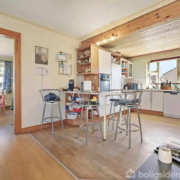 Køkken med barstole og spisebord. Rummet har træloft, lyse skabe, og moderne apparater. I baggrunden ses en spisestue med et vindue og et mønstret dug på bordet.