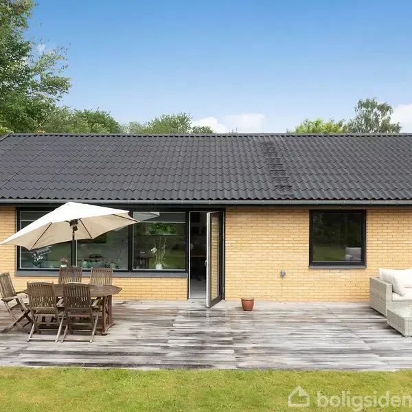 Et gult murstenshus med sort tag står stille i en grøn have. På terrassen er der et bord med stole under en parasol ved siden af en grill og en sofa.