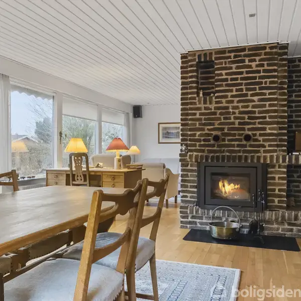Stue med møbler og lamper. To bløde lænestole vender mod en sofa omkring et lavt bord. Store vinduer viser udsigt til en have. Væggene er lyse.