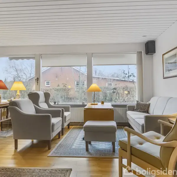 Et murstenshus står i en have med en flisebelagt gangsti og omgivet af grønne buske og træer. Huset har en tagryg og flere vinduer.