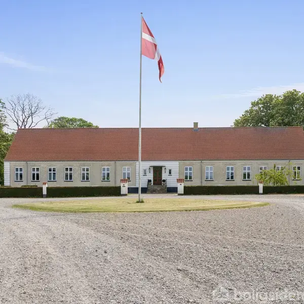 Et langt, gult hus står bag en grusvej med en flagstang foran, omgivet af grønne træer og en blå himmel.