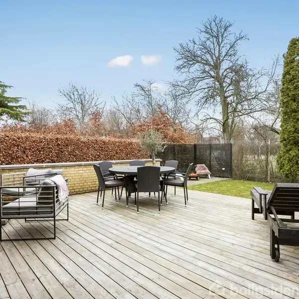 Terrasse med trædæk omfatter en spisegruppe, solsenge og en sofa; omkranset af hække og høje træer, skaber det en hyggelig og privat atmosfære i en haveindstilling.