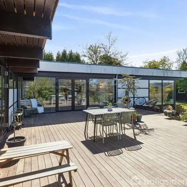Et moderne hus har en stor træterrasse med havemøbler og planter. Terrassen fører til en glasdør, der åbner ind til en stue. Der er en grøn have i baggrunden.