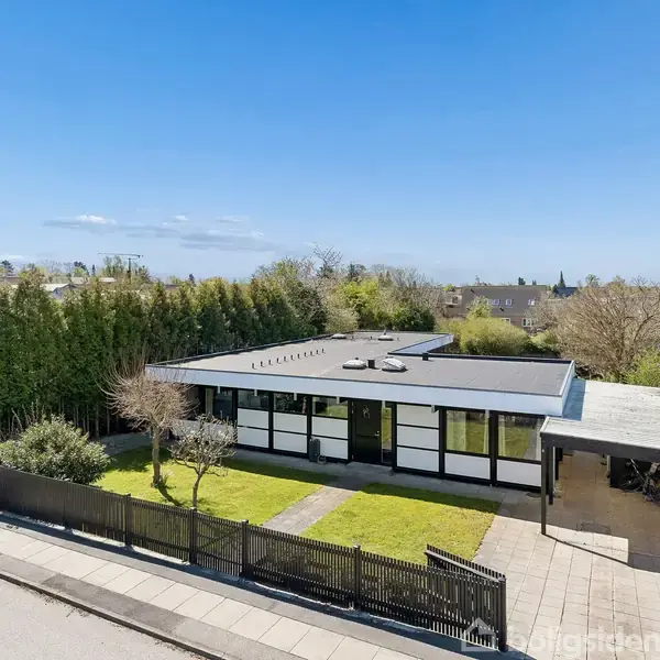 Fladt, moderne hus med sorte og hvide paneler står i en grøn have. Huset har en indgangssti og en carport. Tætklippede buske omgiver ejendommen i et forstadsområde.