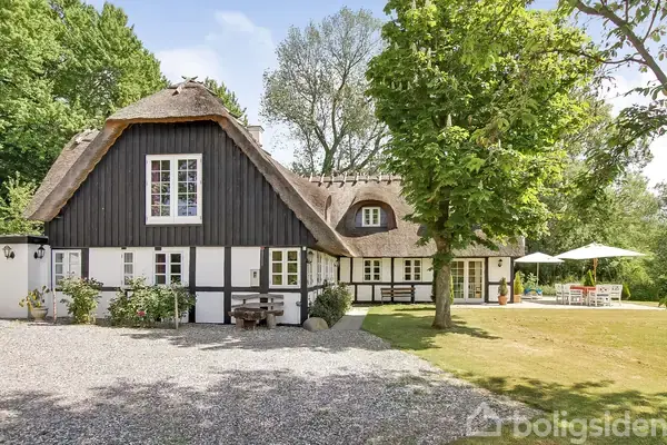 Et bindingsværkshus med stråtag står stille i en grøn have med grussti. Træer omkranser græsplænen, og der er en terrasse med havemøbler i baggrunden.