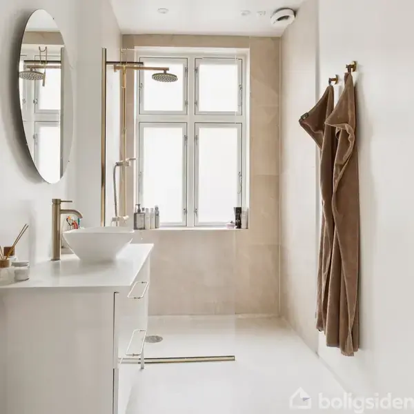 Et moderne badeværelse har en hvid vask med spejl på væggen. Bruseren er ved vinduet, omgivet af beige klinker. Håndklæder hænger på væggen.