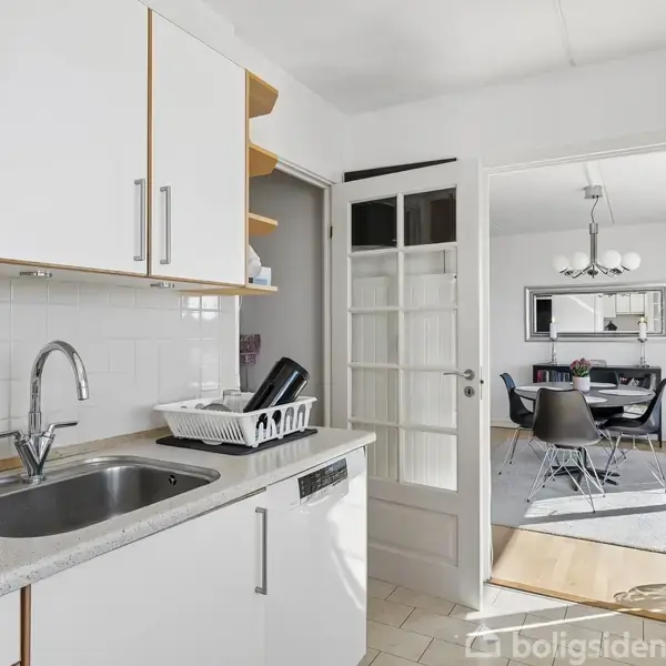 Køkken med hvidt skab og vask, hvor en opvaskestativ står ovenpå. Gennem en glasdør ses en spisestue med bord og stole.