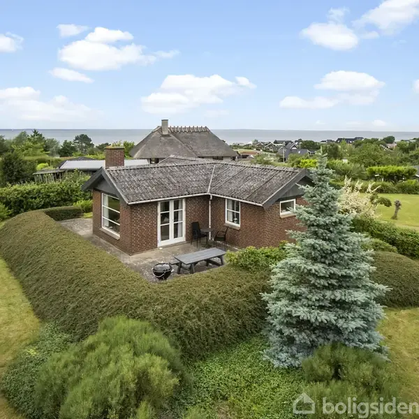 Et lille rødt murstenshus står omgivet af velplejede hække i en grøn have. Der er en terrasse foran huset med havemøbler. Baggrunden viser andre huse og en blå himmel.