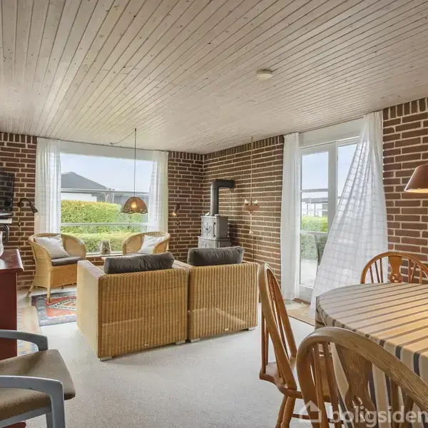 Stue med murstensvægge, sofagruppe foran tv, spisebord med stole i forgrund. Store vinduer og glasdør mod terrasse giver lys. Træloft og tæppebelagt gulv skaber en hyggelig atmosfære.