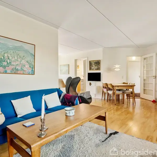 Blå sofa står langs væggen med et maleri hængende over. Træbord foran sofaen. Spisestue med træbord og stole i baggrunden. Trægulve og hvidmalede vægge skaber lys og rummelig atmosfære.
