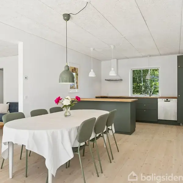 Spisebord med hvid dug og blomster står i lys stue. I baggrunden ses et moderne køkken med grå skabe og vindue, omgivet af lyse vægge og trægulv.