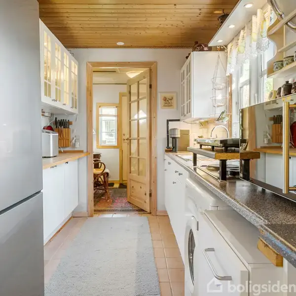 Et smalt køkken med en stålgrå køleskab, hvide skabe, og en lang grå bordplade. En glasdør fører til et tilstødende rum. Træloft og flisegulv kompletterer rummet.