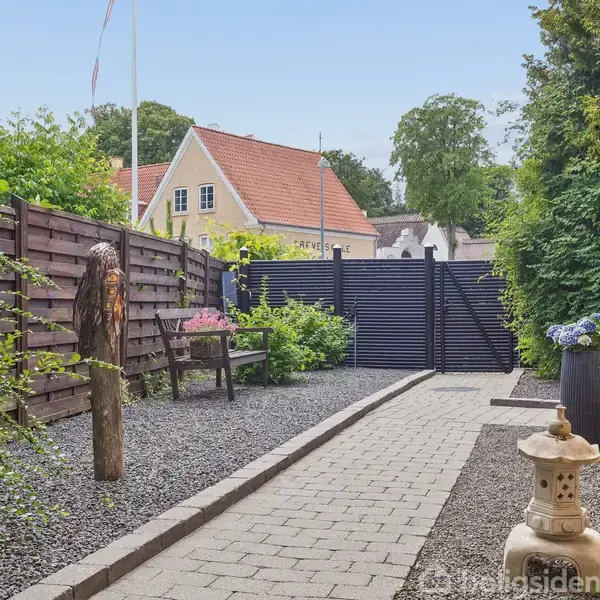 Gangen fører til en lille have med grussti flankeret af træhegn og buske. En stenterrasse har en japansk stenlanterne og en stor plantekrukke til højre.