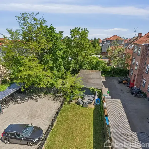 En balkon med trægulv har en glasgelænder ved en murstensvæg. En dør fører indad. Udsigten inkluderer grønne træer og blå himmel.