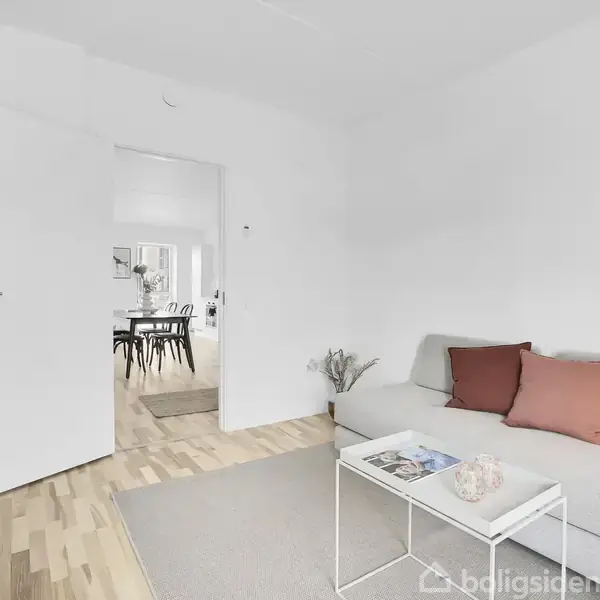 En grå sofa med røde puder står i en lys stue. Et lille hvidt bord med magasiner foran sofaen. Åben dør fører til en spisestue med sort-hvide stole.