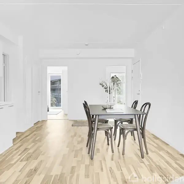 Et spisebord med fem stole står i et lyst køkken-alrum. Bordet har en vase med blomster. Til venstre er der en hvid køkkenvæg med skabe og vask.