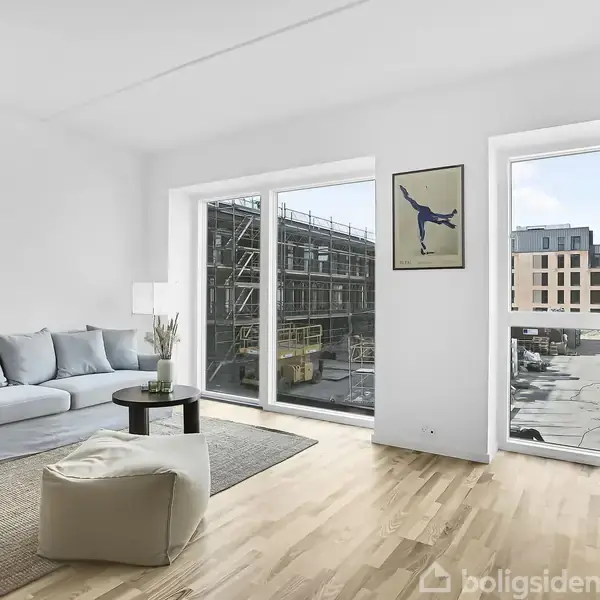 En lys stue med en blå sofa og et lille bord på et tæppe. Store vinduer viser en byggeplads udenfor. En plakat hænger på væggen ved siden af vinduet.