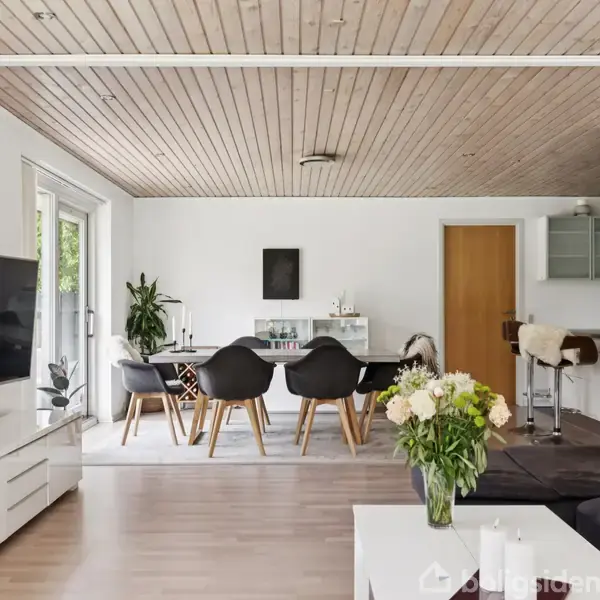 Stue med en stor sofa, et lavt sofabord fyldt med blomsterbuketter, en fladskærms-TV på væggen, og et spisebord med sorte stole i en stilfuld, moderne indretning.