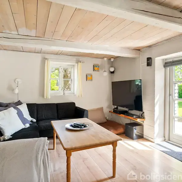 Sort hjørnesofa med puder står i en lys stue. Træbord foran sofaen. Fladskærms-TV placeret i hjørnet. Vinduer og dør slipper sollys ind og viser have udenfor.