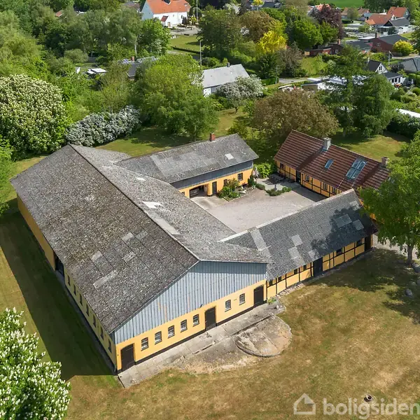 En stor, L-formet gård med gult murværk og gråt tag ligger i et frodigt grønt område med træer. Omkringliggende huse er synlige i baggrunden.