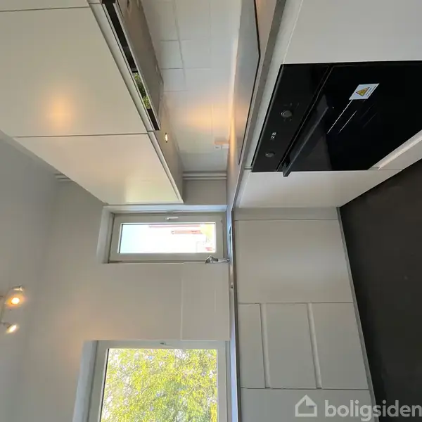 Hvidt køkken med indbygningsovn og skabsløsninger. Et vindue med udsigt til træ og dagslys. Moderne armaturer og sort gulv fuldender det minimalistiske design.